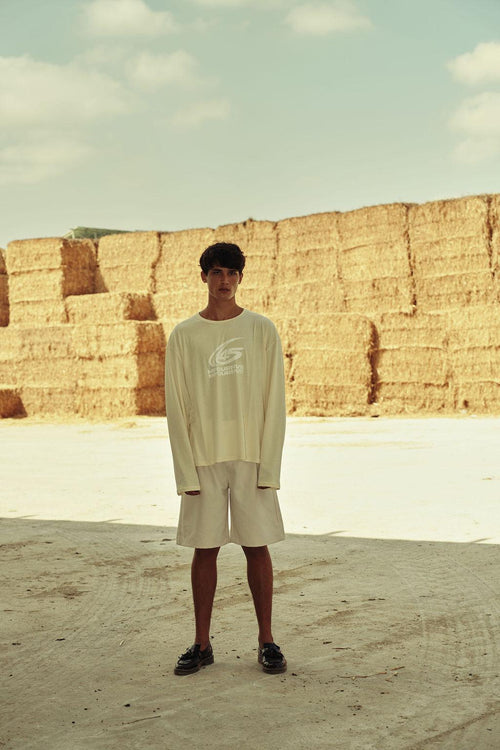 Full-body shot of male model in cream long-sleeve graphic t-shirt and shorts in front of hay bales