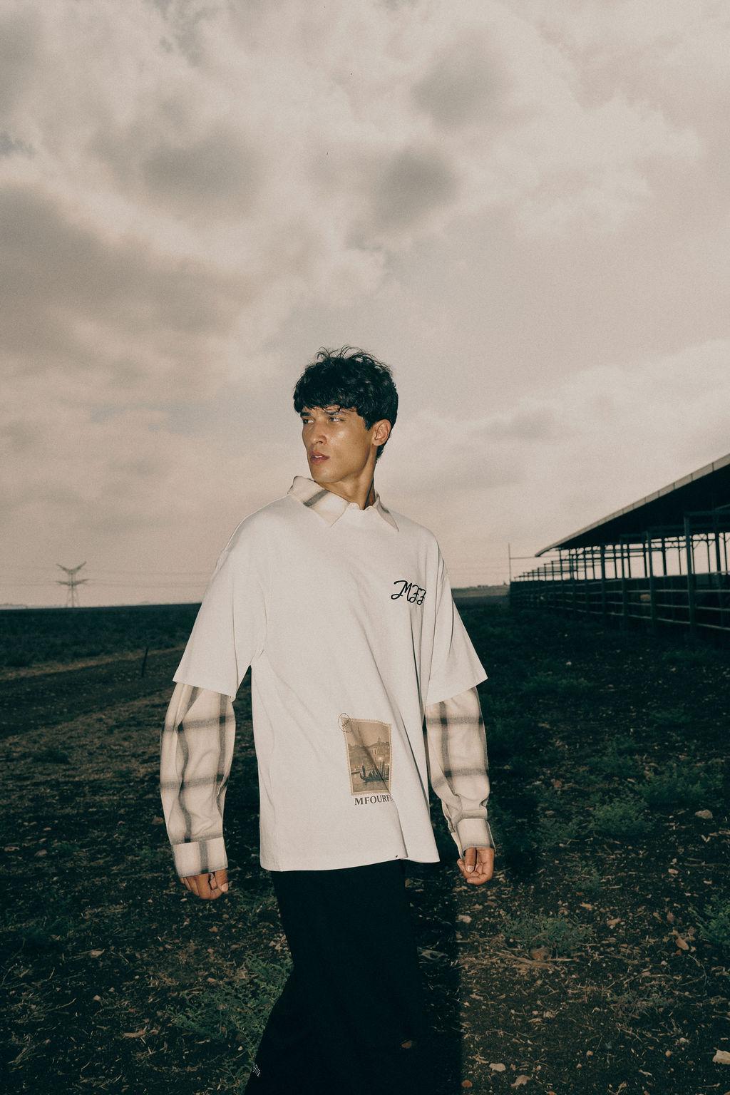 Male model wearing white graphic t-shirt layered over a plaid long-sleeve shirt outdoors
